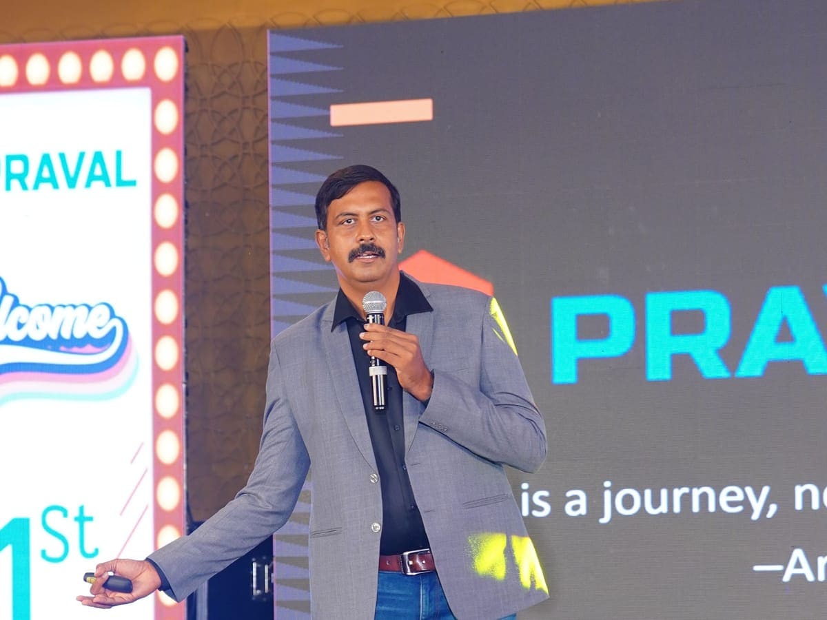 A Praval leader speaking on stage with a microphone, in front of the Praval screen